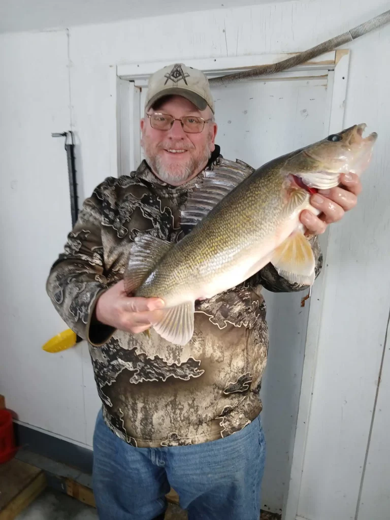 big walleye catch river bend fishing report Man in camouflage holding a large walleye against a white wall.