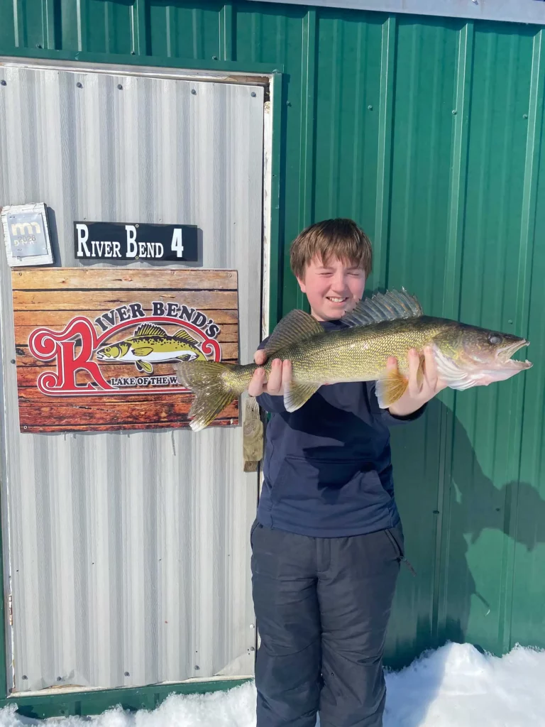 river bend resort ice house walleye fishing Young angler holding a walleye in front of a green wall with a River Bend Resort sign in an ice house.